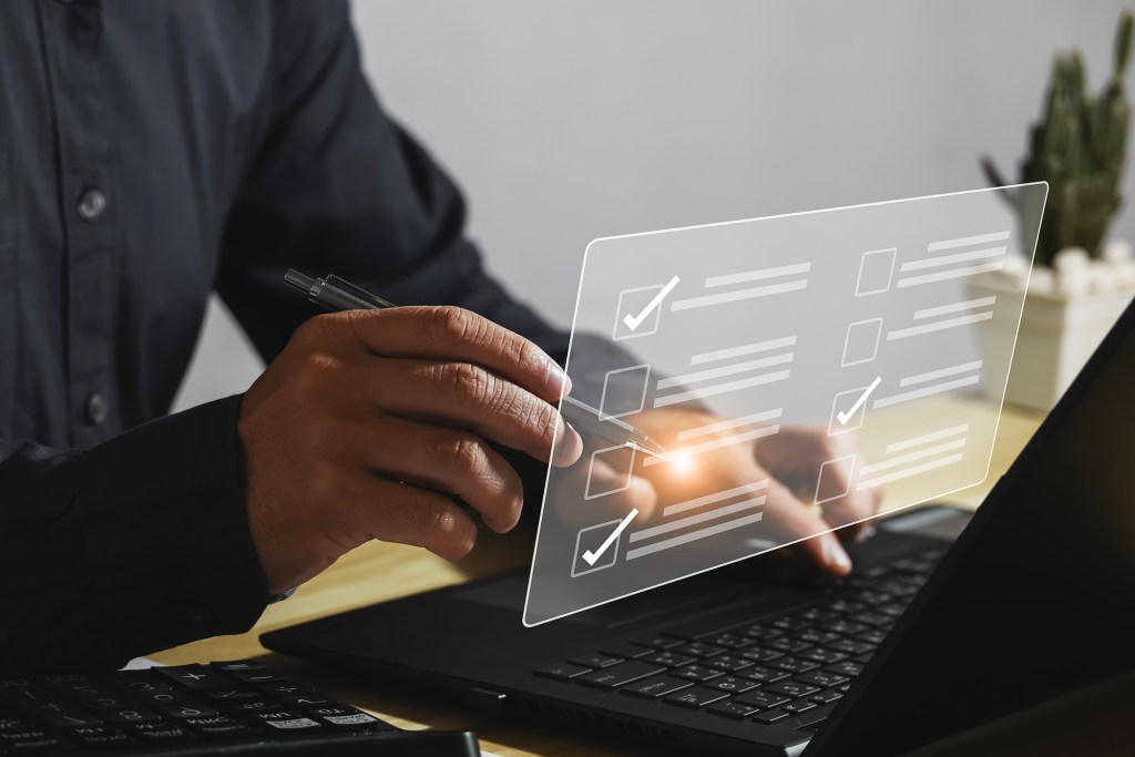 A person using a stylus to interact with a digital checklist displayed over a laptop, symbolizing survey design and data collection.