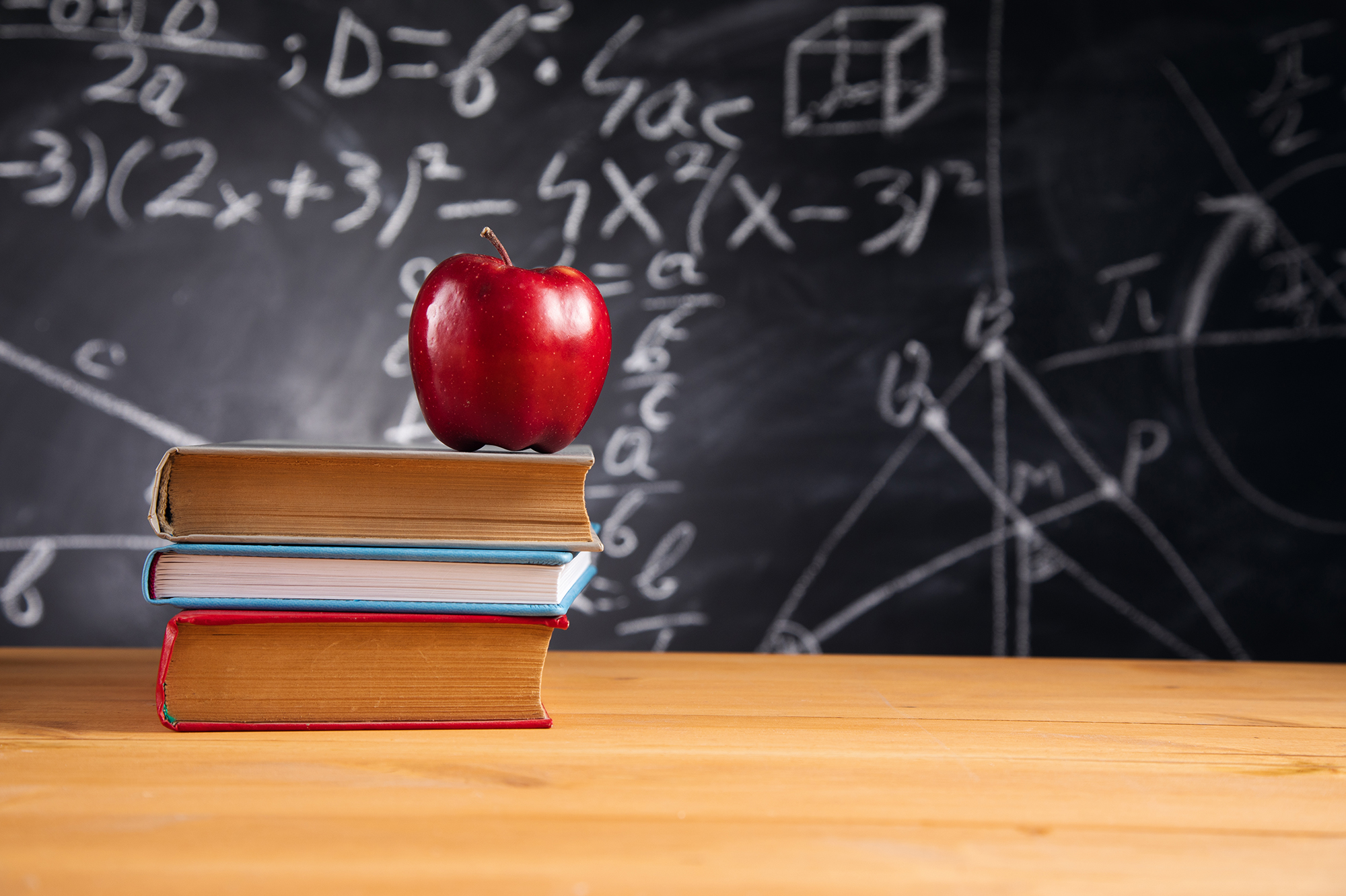 A red apple sitting on top of three stacked books in front of a chalkboard covered with math equations and diagrams.